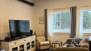 A hotel room with a bed, a sofa, a chair, a TV, a small table, and a window with curtains. - Serene Studio overlooking the river (Juneau)