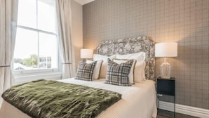 Una cama bien hecha con una manta verde, dos lámparas de noche y un cabecero estampado con motivos florales.