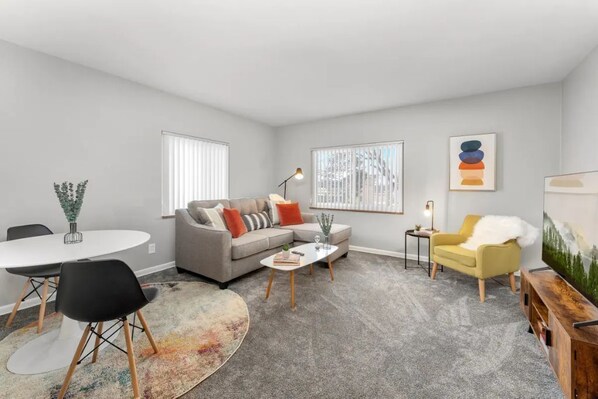 A modern living room with a grey sofa, a yellow armchair, a white dining table, and a coffee table. There are two windows with blinds, a painting on the wall, and a TV on a wooden stand.