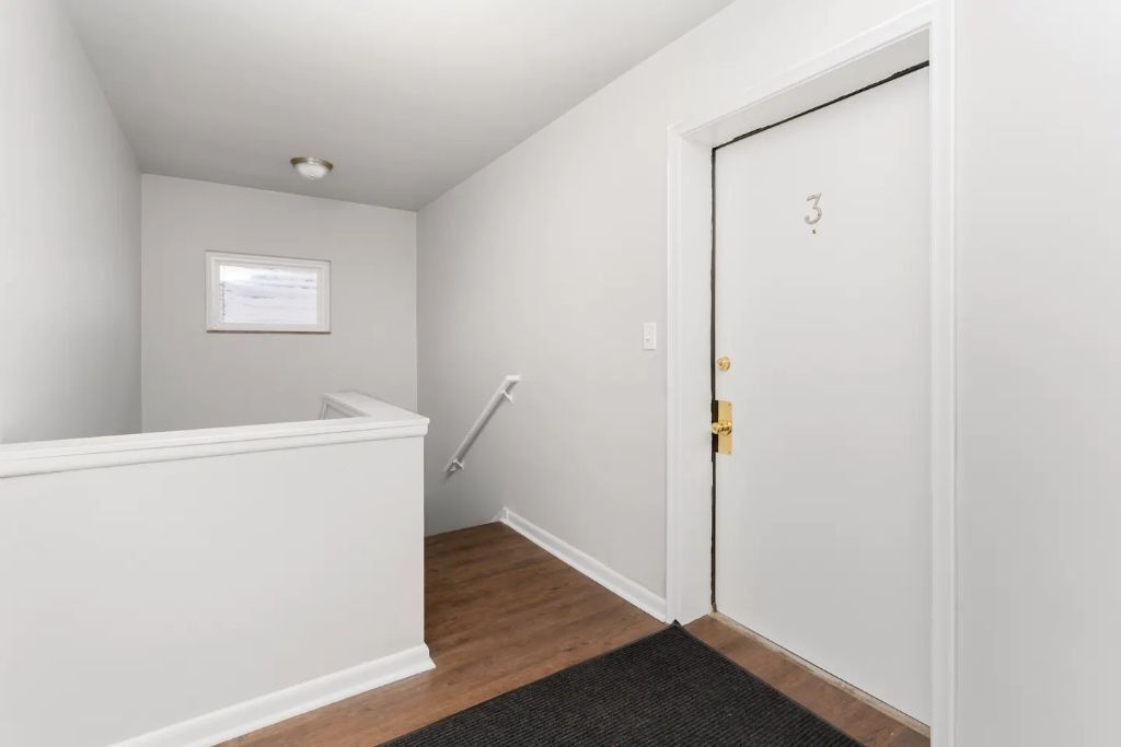 A narrow hallway with white walls, a wooden floor, a black doormat, a white door with a number, and a small window.