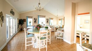 Une salle à manger spacieuse avec une table ronde, des chaises et un lustre. On y trouve plusieurs fenêtres avec des rideaux, un ventilateur de plafond et une table à manger agrémentée d’un vase de fleurs.