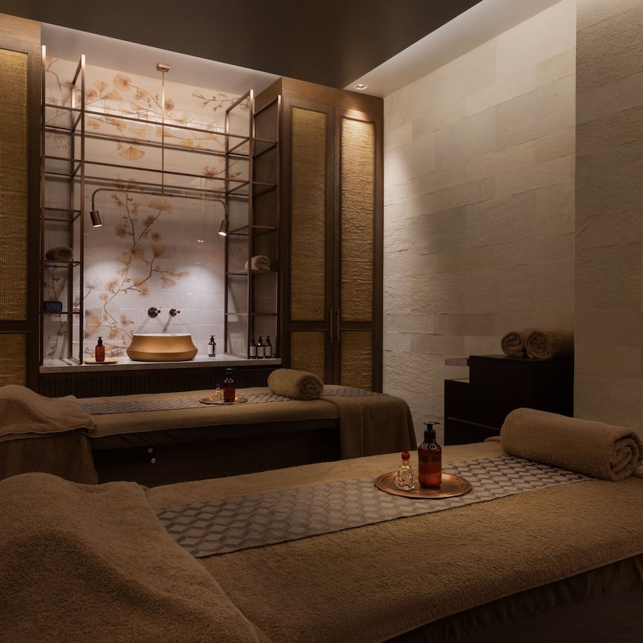 A spa room with two massage tables, a wall-mounted shelf with toiletries, and a decorative wall panel.