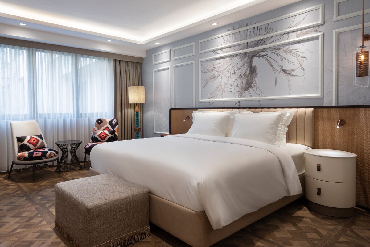 A hotel room with a large bed, a side table, a chair, a small table, and a large window with curtains.