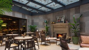 A spacious indoor area with round tables and chairs, a fireplace, and a large window with a view of the sky. - Burdock Hotel Istanbul, Autograph Collection (Istanbul)