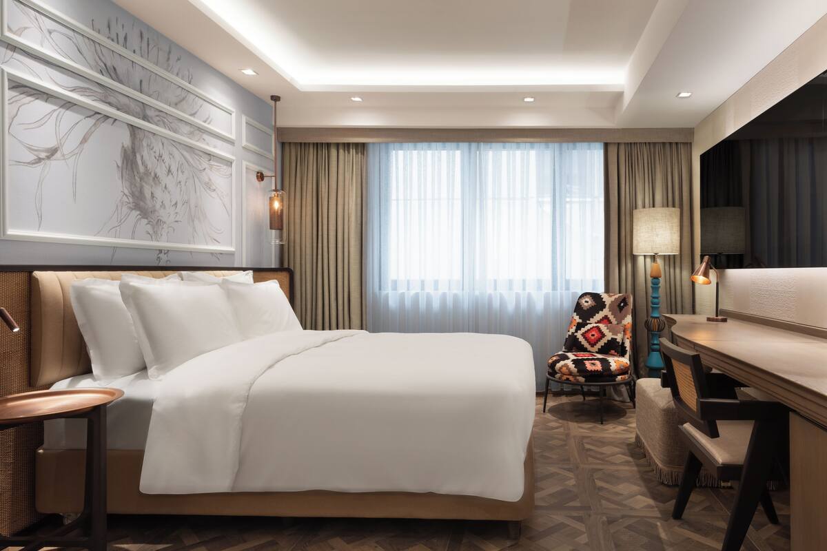 A modern hotel room with a large bed, a desk, and a chair. There is a large window with curtains, a wall-mounted artwork, and a lamp on the desk.