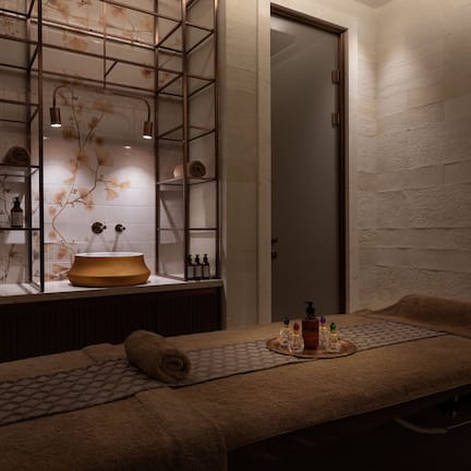 A spa room with a massage table, a wooden basin, and shelves with bottles and a lamp.