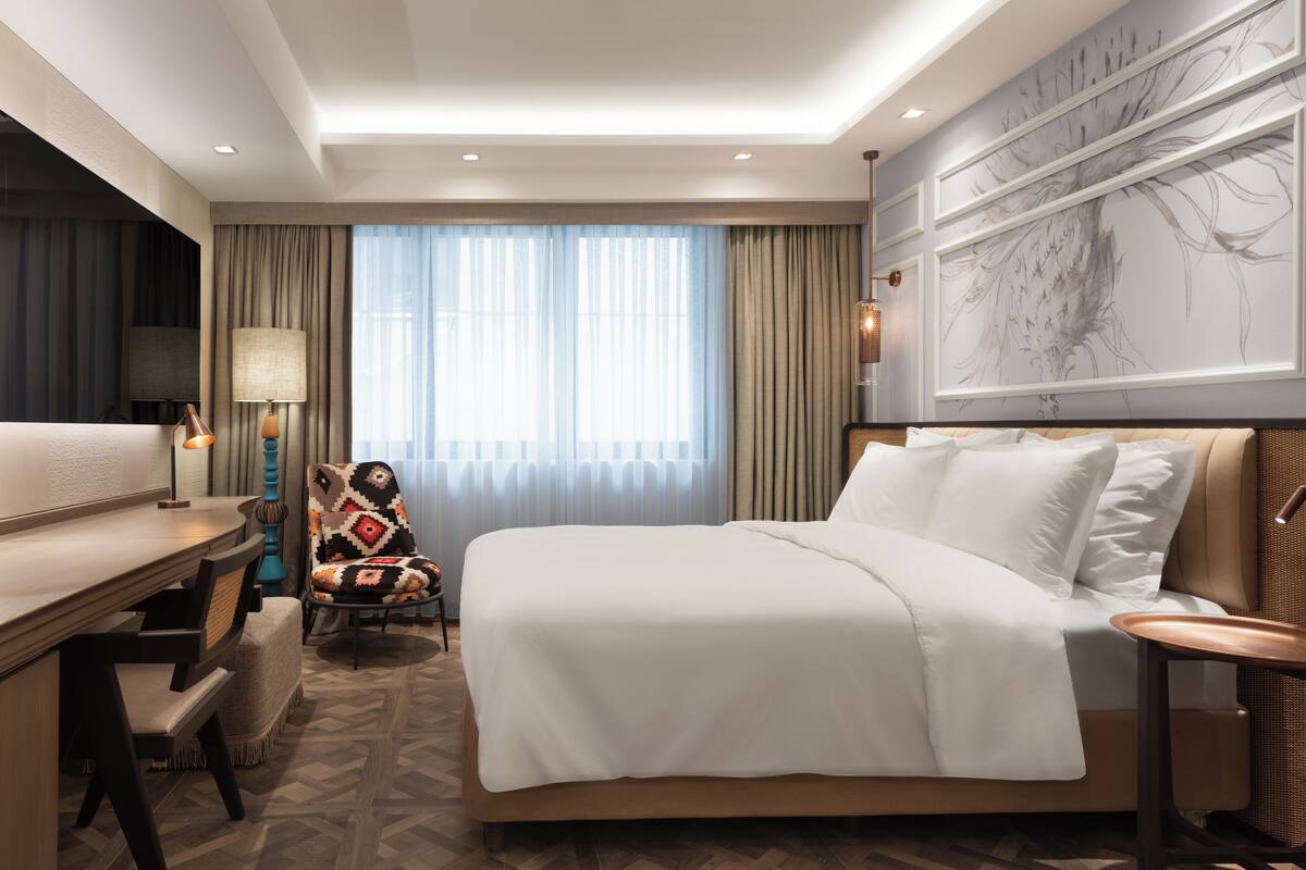 Hotelkamer met een groot bed, een bureau met lamp, een stoel en een groot raam met gordijnen.