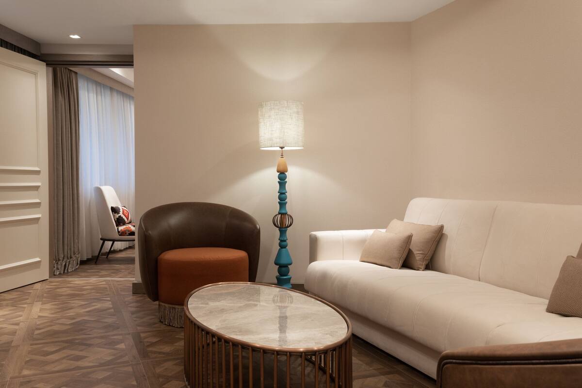 A modern hotel room with a beige sofa, a brown leather armchair, a round coffee table, and a tall floor lamp.