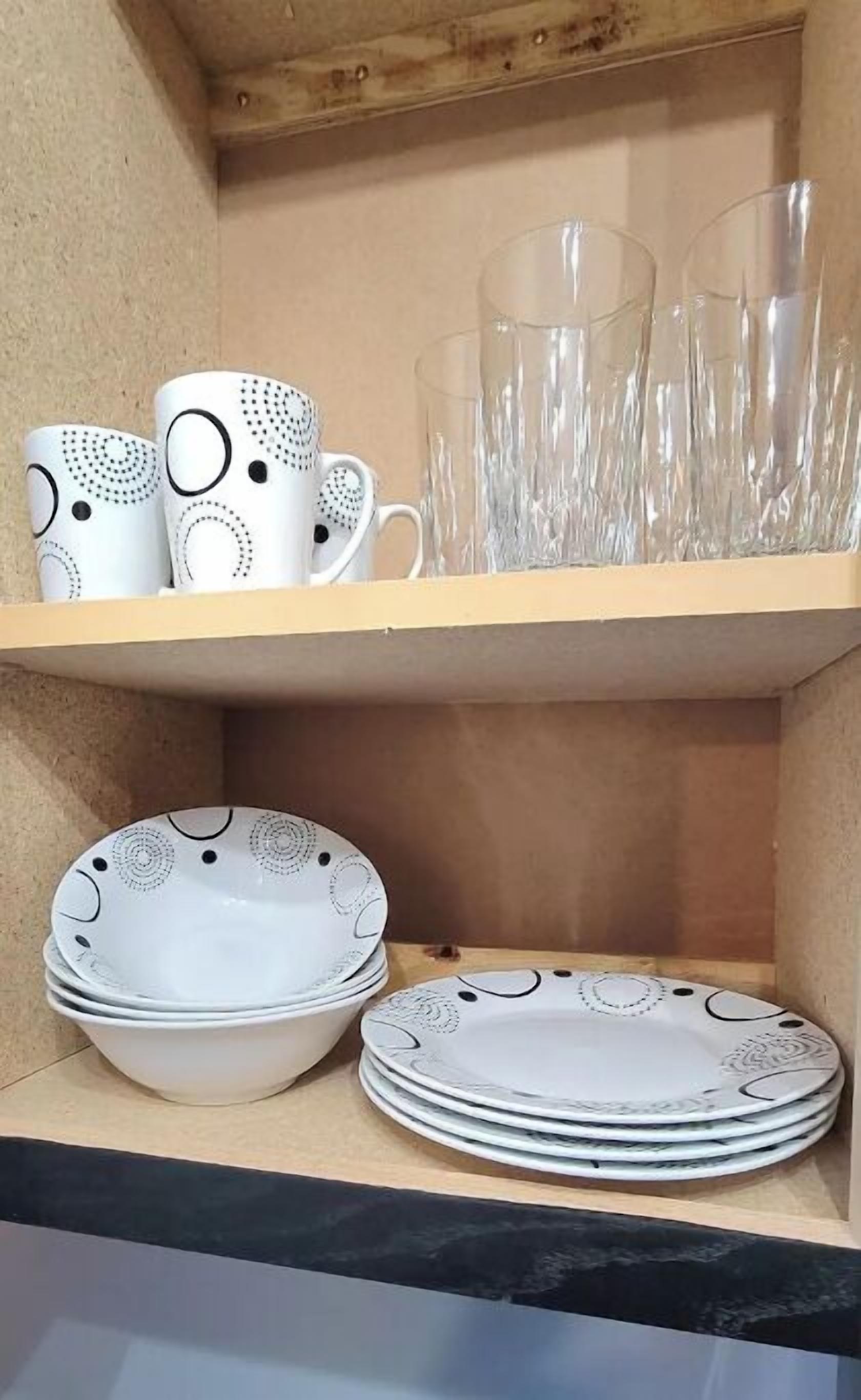 Une étagère avec des tasses en céramique blanche, des verres à boire transparents et des assiettes blanches ornées de motifs noirs.