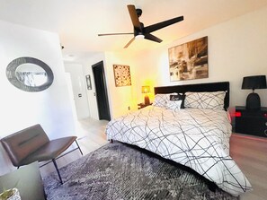 A bedroom with a bed, a ceiling fan, a desk, a chair, a lamp, and a painting on the wall. - Tuxedo Touch Modern 2BR Townhouse (Smyrna)