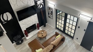 A modern living room with a fireplace, a brown leather sofa, a round coffee table, and a large window with a view of the outdoors. - Tuxedo Touch Modern 2BR Townhouse (Smyrna)