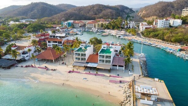An aerial view of a coastal town with a marina, beach, and buildings with balconies overlooking the water.
