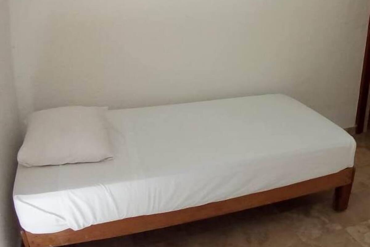 A single bed with a wooden frame and white bedding in a room with plain walls and a tiled floor.