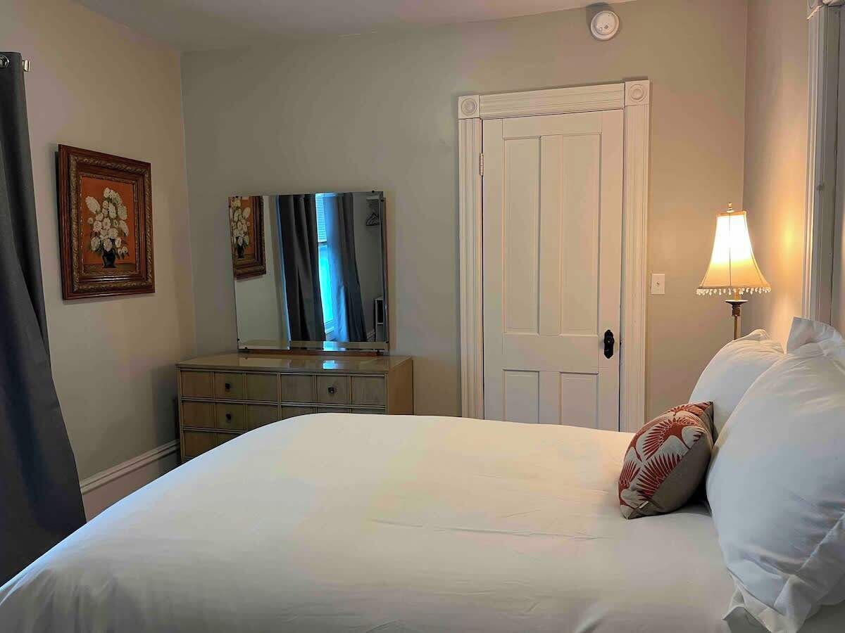 Una cama bien tendida con sábanas blancas y una única almohada decorativa, un aparador con espejo y una lámpara en la mesita de noche.
