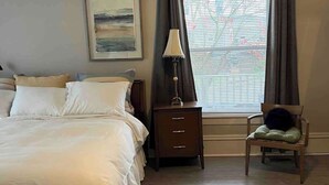 Un dormitorio con una cama grande, una mesita de noche, una silla, una lámpara, una ventana con cortinas y un ventilador de techo.