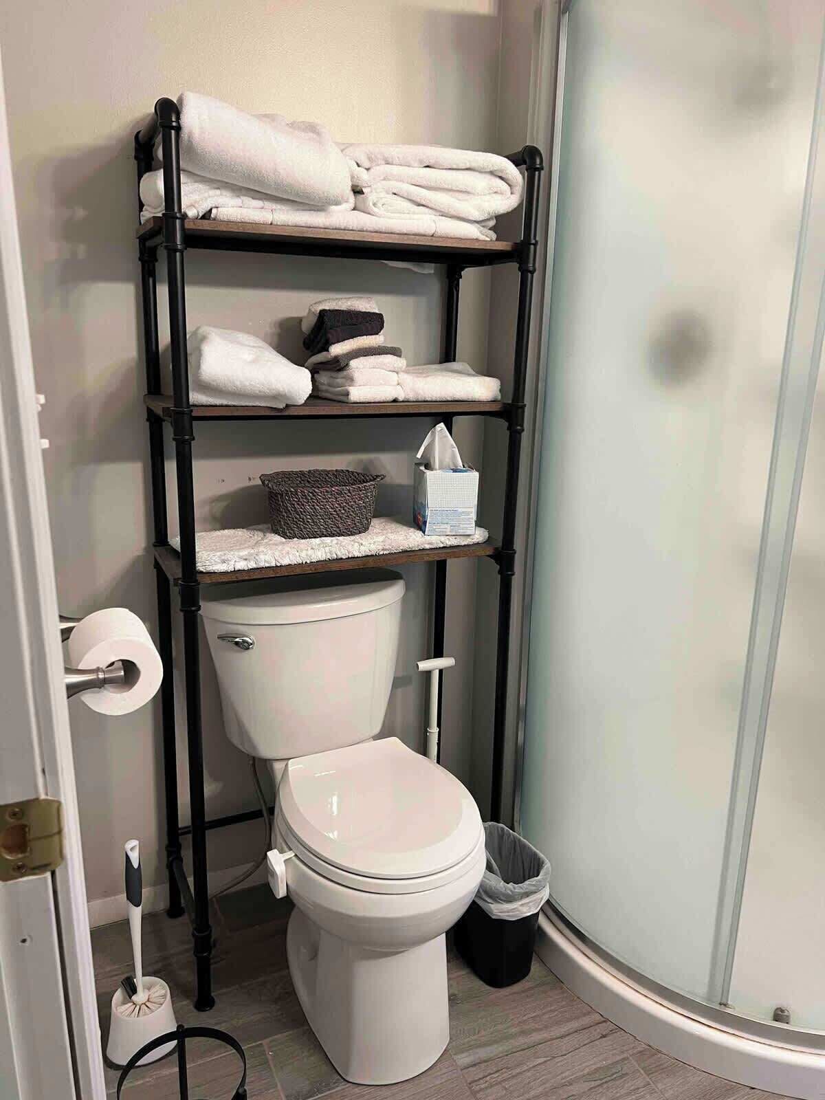 A bathroom with a white toilet, a black shelf unit with towels, a basket, and a toilet paper holder.