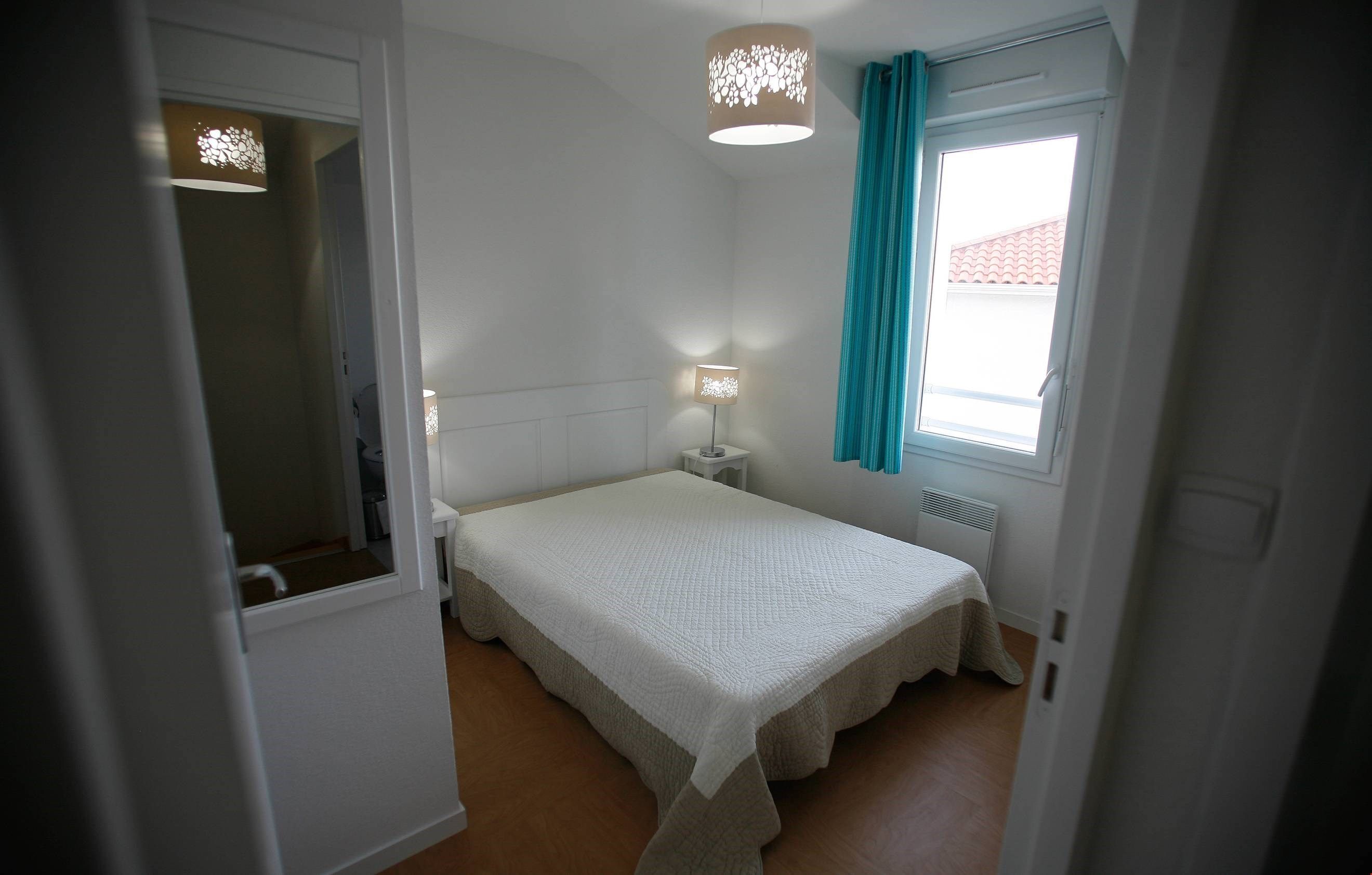 A bedroom with a single bed, a bedside table with a lamp, a window with curtains, and a radiator.