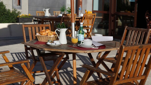 An outdoor dining area with wooden tables and chairs, a basket of croissants, a pitcher, a bottle of water, and a cup of coffee.