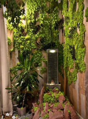 Espacio interior estrecho con paredes verticales cubiertas de exuberantes enredaderas, un pilar central con una fuente de luz y una planta en maceta.