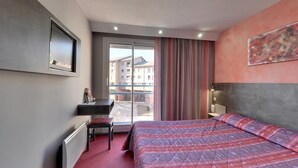A hotel room with a bed, a desk, a chair, a radiator, a TV mounted on the wall, and a window with curtains.