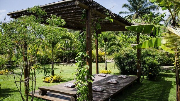 A wooden deck with cushions, surrounded by lush greenery and a pergola structure. - Pousada Apoema (Maraú)