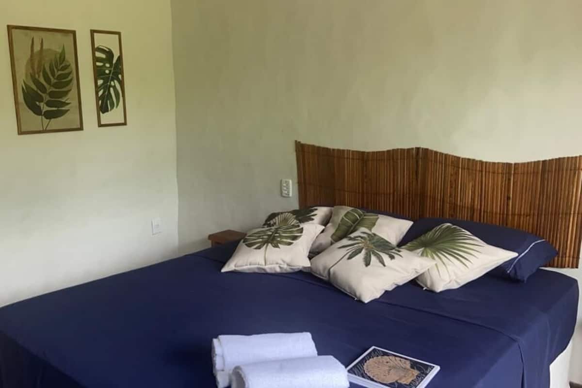 Un lit recouvert d’une courtepointe bleu foncé, plusieurs oreillers ornés de motifs de feuilles tropicales et une serviette soigneusement pliée posée sur le lit.