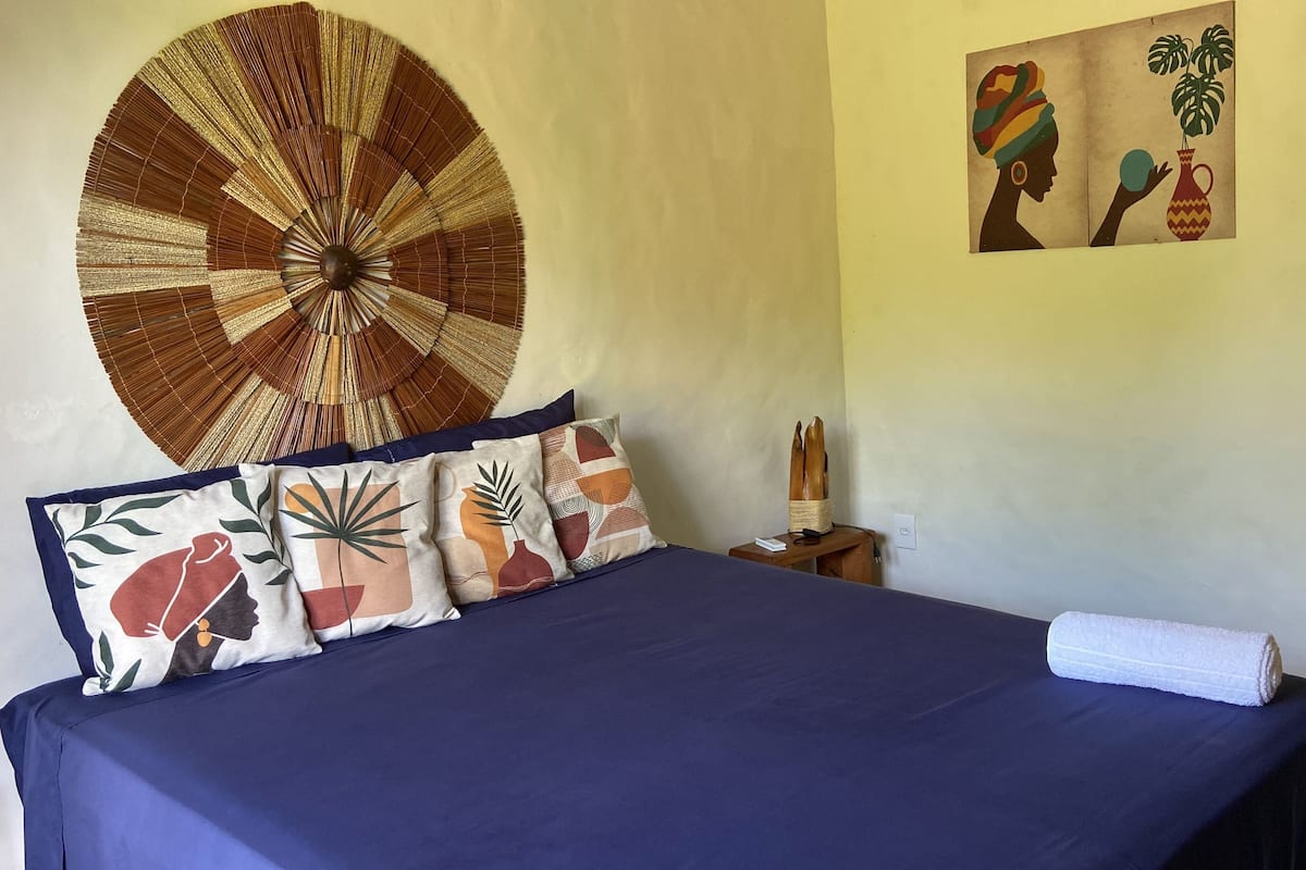 Un lit recouvert d’une couvre-lit bleue, plusieurs coussins ornés de motifs végétaux et géométriques, une grande tapisserie murale tissée et une œuvre d’art fixée au mur représentant une femme et une plante.