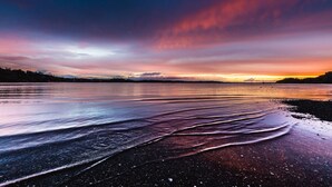 A beach with wet sand and gentle waves, a sunset in the background, and a distant shoreline. - Seaside on Waiheke - Walk to Oneroa Village (Waiheke Island)