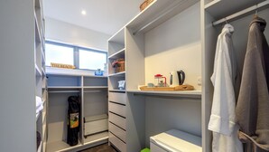 A well-organized closet with shelves, drawers, and hanging space, a small trash bin, and a window with a view of the beach. - Seaside on Waiheke - Walk to Oneroa Village (Waiheke Island)