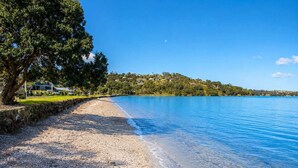 A beach with a stone wall, clear blue water, and a tree-lined shore. - Seaside on Waiheke - Walk to Oneroa Village (Waiheke Island)