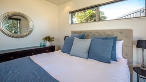 A bedroom with a large bed, a round mirror, a dresser, a nightstand, and a window with a view of trees. - Seaside on Waiheke - Walk to Oneroa Village (Waiheke Island)