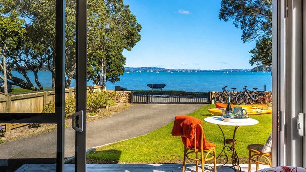 A view from a room with a glass door to a deck, overlooking a body of water with boats and a distant shoreline. - Seaside on Waiheke - Walk to Oneroa Village (Waiheke Island)