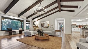 A spacious living room with a high ceiling, wooden beams, a large window, and a view of the outdoors.