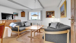 A cozy living room with a wooden table, two grey sofas, a television, and a window with curtains.