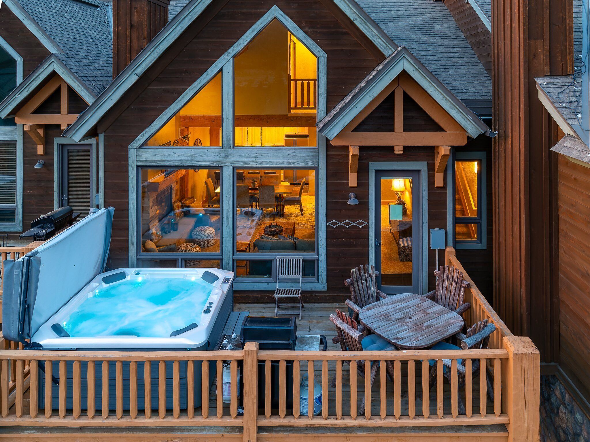A wooden cabin with a hot tub on the deck, large windows, and a cozy interior.