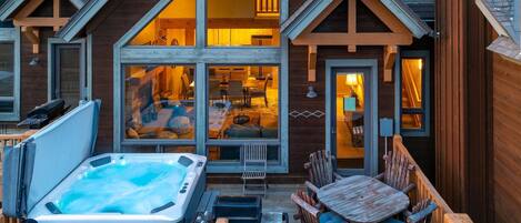 A wooden cabin with a hot tub on the deck, large windows, and a cozy interior.