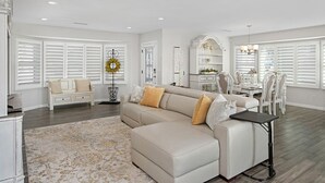 Un salon spacieux avec un canapé d’angle beige, un coin repas avec des meubles blancs et de grandes fenêtres dotées de volets blancs.