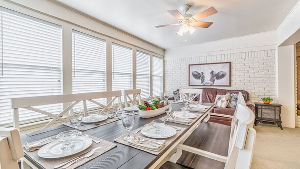 A dining area with a long table set for six, white brick walls, and a ceiling fan.