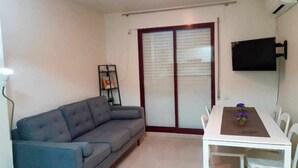 Un soggiorno con un divano grigio, un tavolo da pranzo bianco con sedie, una TV a schermo piatto montata a parete e una porta-vetro scorrevole che dà su un balcone.