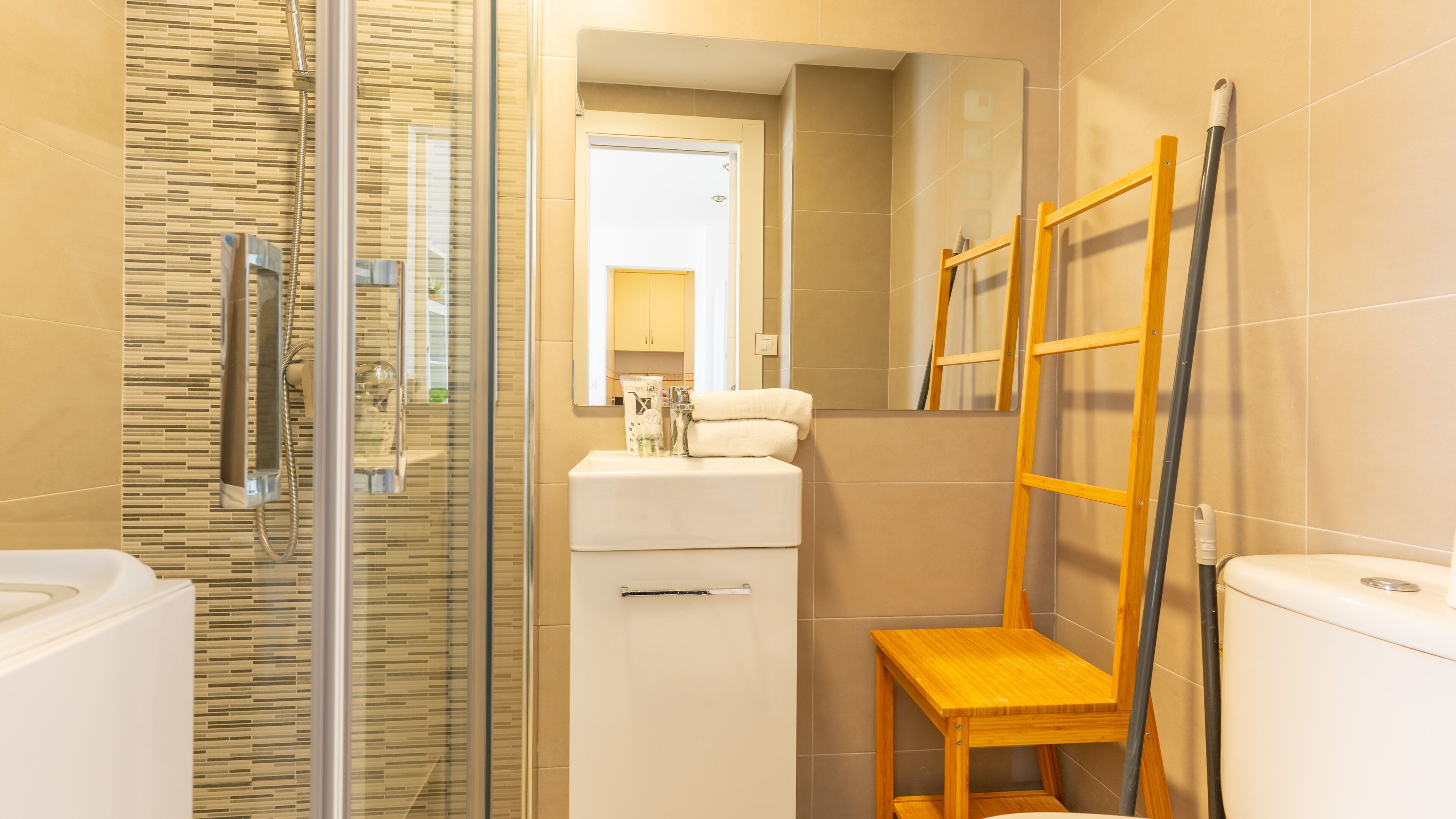 A bathroom with a glass shower enclosure, a white sink, a wooden chair, and a toilet.