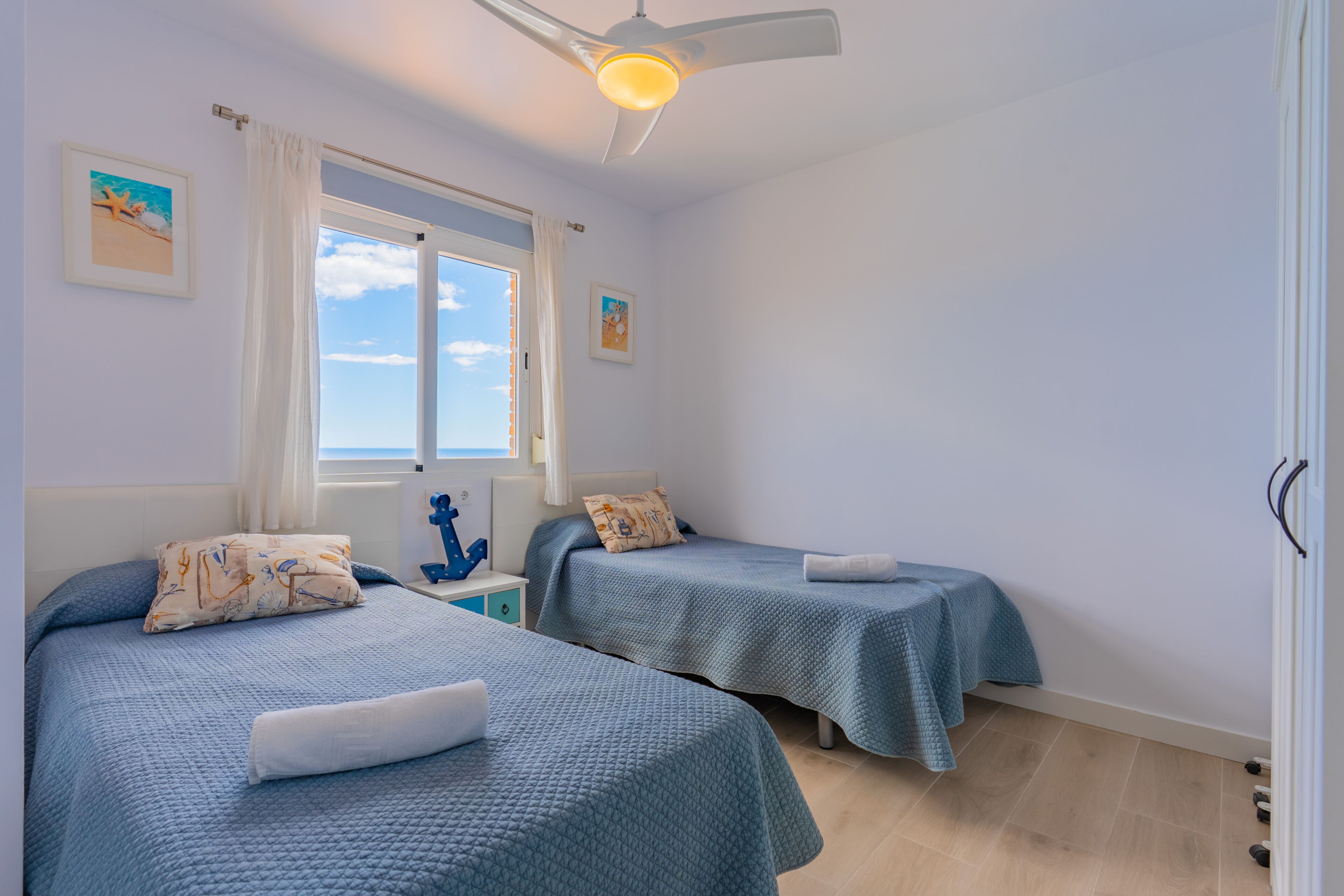 A bedroom with two beds, a ceiling fan, a window with curtains, and a small nightstand with a decorative item.
