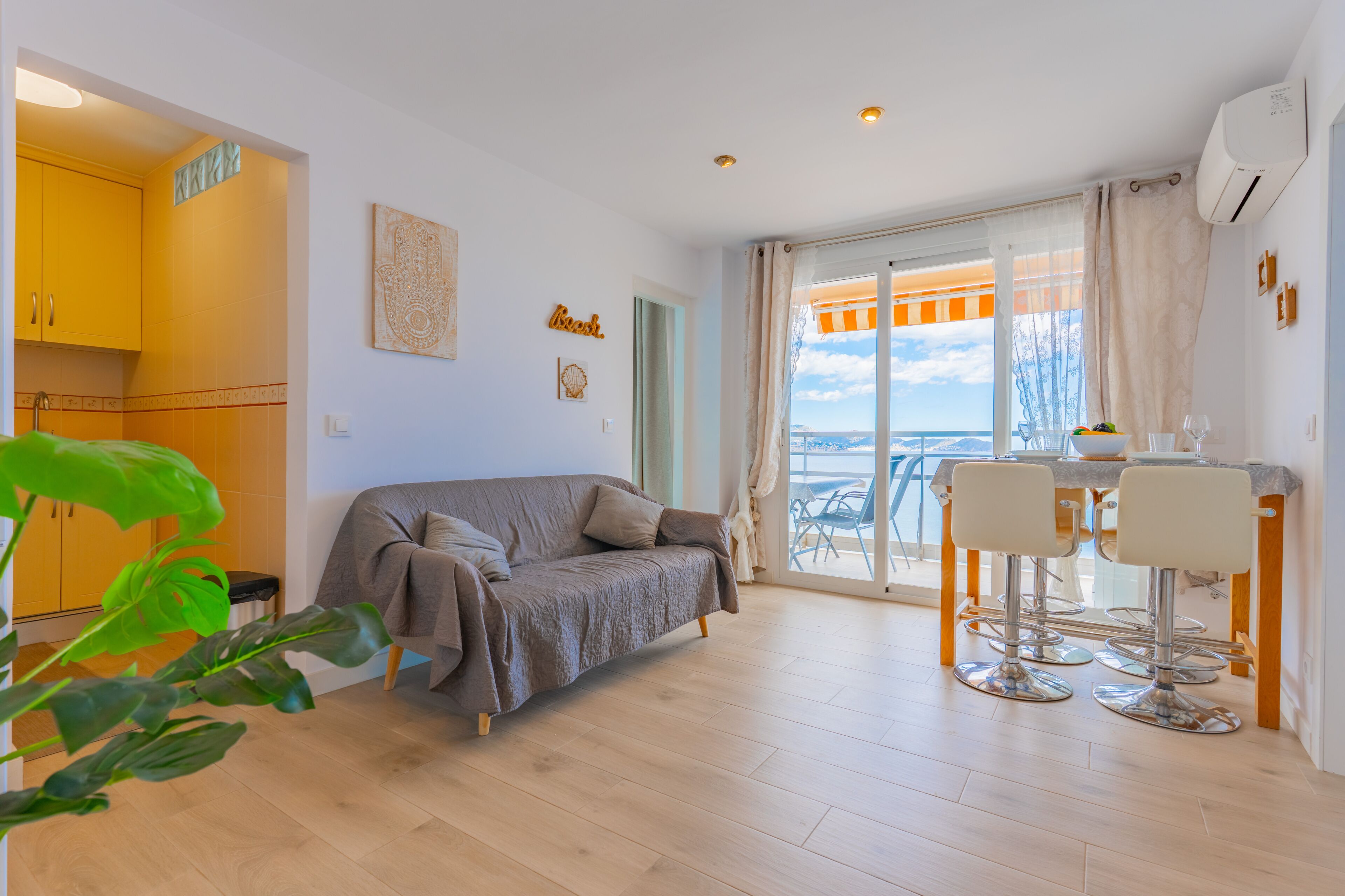 A modern living room with a sofa, a dining area with chairs, a view of the sea, and a kitchen with yellow cabinets.