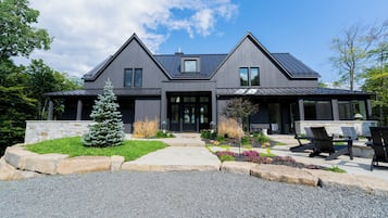 Une maison moderne à deux étages, avec des fondations en pierre, un toit noir et un jardin bien entretenu à l’avant, agrémenté d’un petit arbre et de parterres de fleurs.