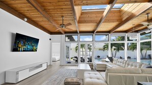 A modern living room with a large TV, a sofa, a coffee table, and a view of a pool and outdoor area. - 2260 Managed By Brampton Park (Pompano Beach)
