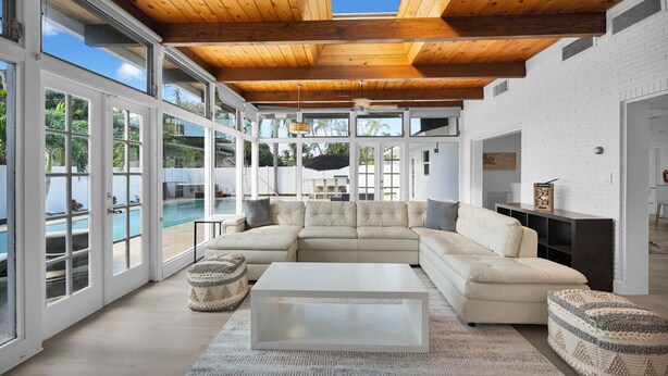 A spacious living room with a large sectional sofa, a coffee table, and a view of a swimming pool through large windows. - 2260 Managed By Brampton Park (Pompano Beach)