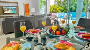 Desayuno montado para dos personas, con platos de croissants, fruta y bebidas, y vista a una piscina y palmeras.
