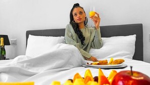 Eine Person sitzt auf einem Bett und isst von einem Teller, neben einem Glas Orangensaft.