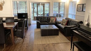 A living room with a sofa, ottoman, and a piano. There is a television and a dining table with chairs. - La Maison jaune: Spa, comfort, wifi (Chute-Saint-Philippe)
