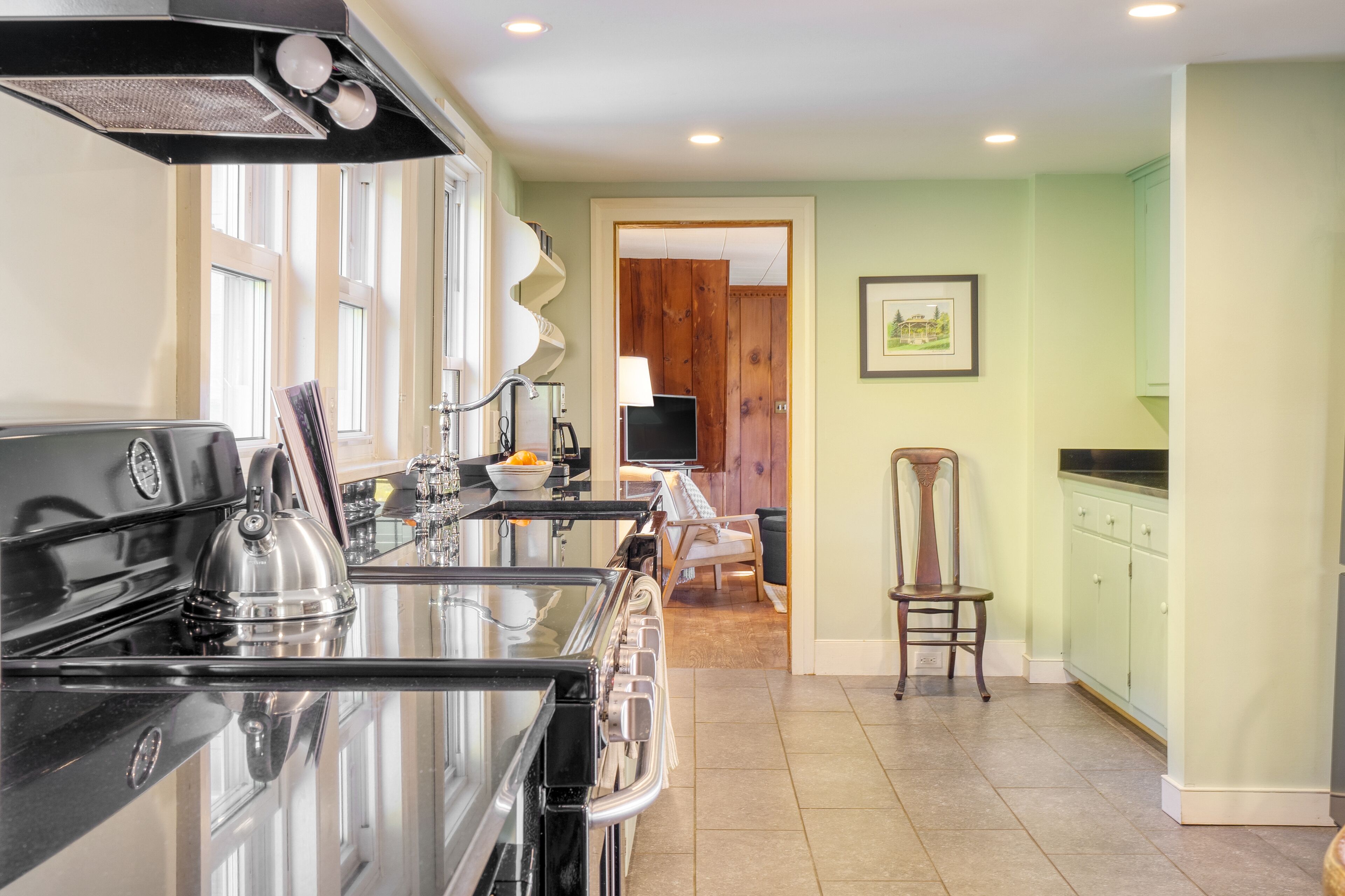 Una cocina con un horno negro, un hervidor y un lavabo. Se ve una silla y una puerta de madera al fondo.