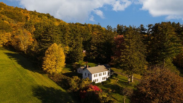 Una casa blanca con fachada simétrica, rodeada por un paisaje verde y exuberante con árboles y una colina al fondo.